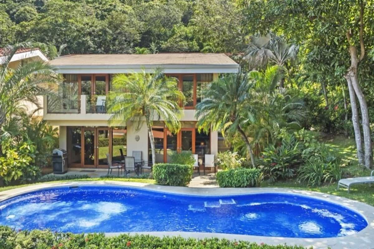 Ein zweistöckiges Haus mit Swimmingpool im Vordergrund, umgeben von üppiger Vegetation und Palmen.