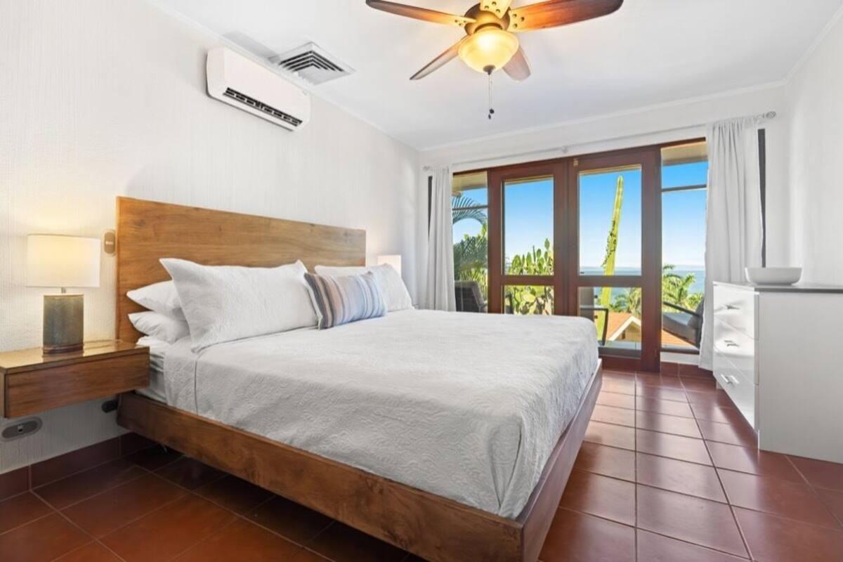 Ein Schlafzimmer mit einem hölzernen Bett, einem Deckenventilator, einer Klimaanlage und Blick ins Freie durch eine Schiebetür.