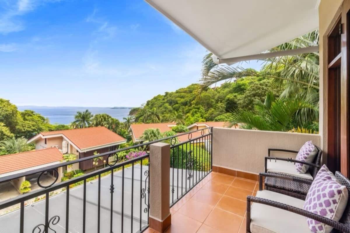 Ein Balkon mit Blick auf eine Küstenlandschaft, darunter Häuser und viel Grün.