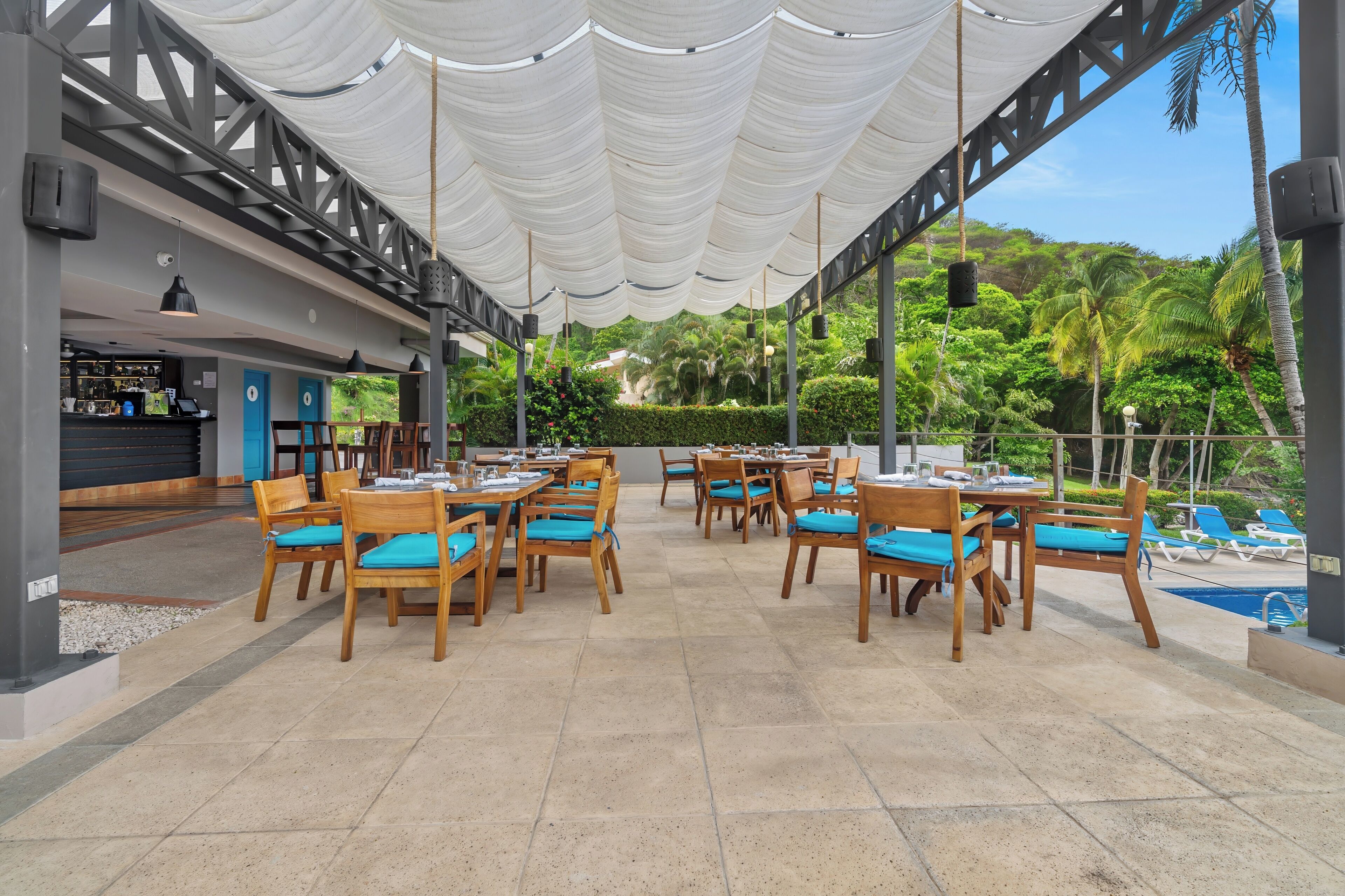 Un área de comedor al aire libre con mesas y sillas de madera, una barra y vista a la piscina.