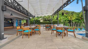 Un área de comedor al aire libre con mesas y sillas de madera, una barra y vista a la piscina.