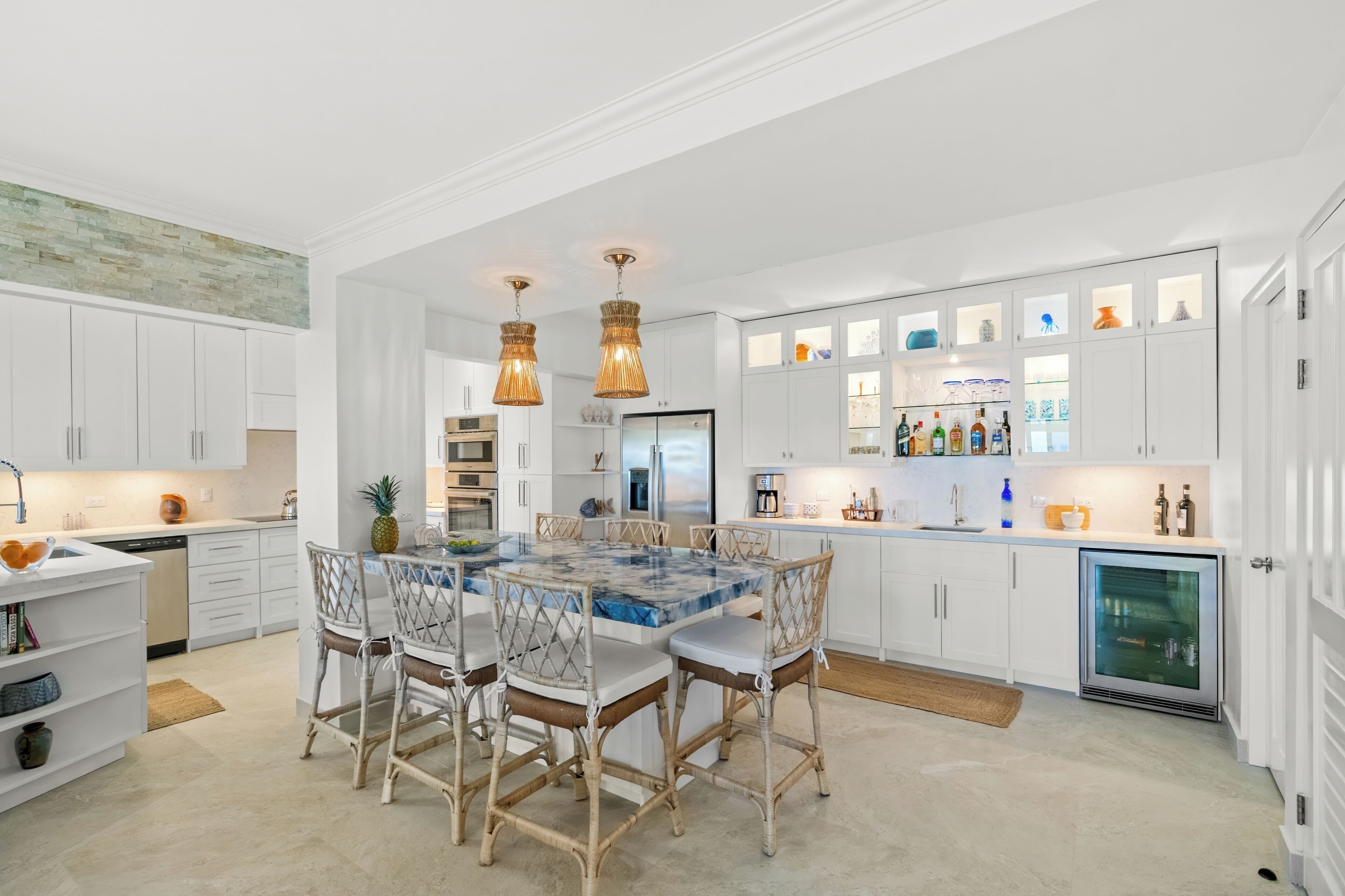 A modern kitchen with white cabinetry, a marble island, wicker chairs, pendant lighting, and a built-in wine cooler.
