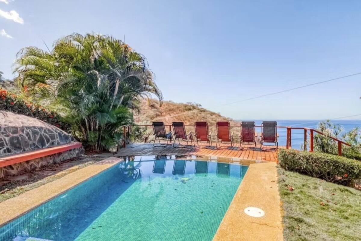 Ein klarer Pool mit Holzterrasse und Liegestühlen, umgeben von tropischer Vegetation und Blick auf den Ozean.