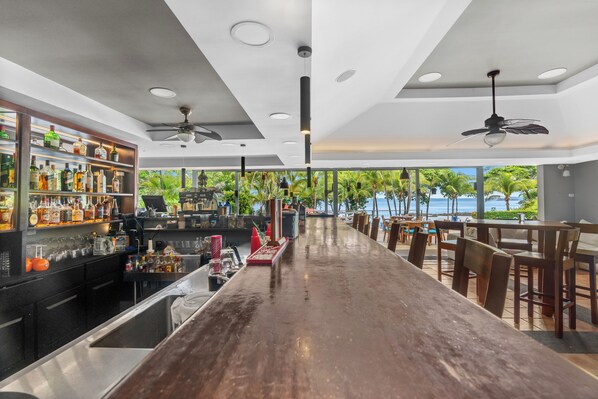 Una zona de bar con una barra larga, estantes llenos de botellas y vista al océano.