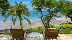 Une vue sur la plage avec deux chaises en osier et une petite table, des palmiers et un ciel bleu dégagé.