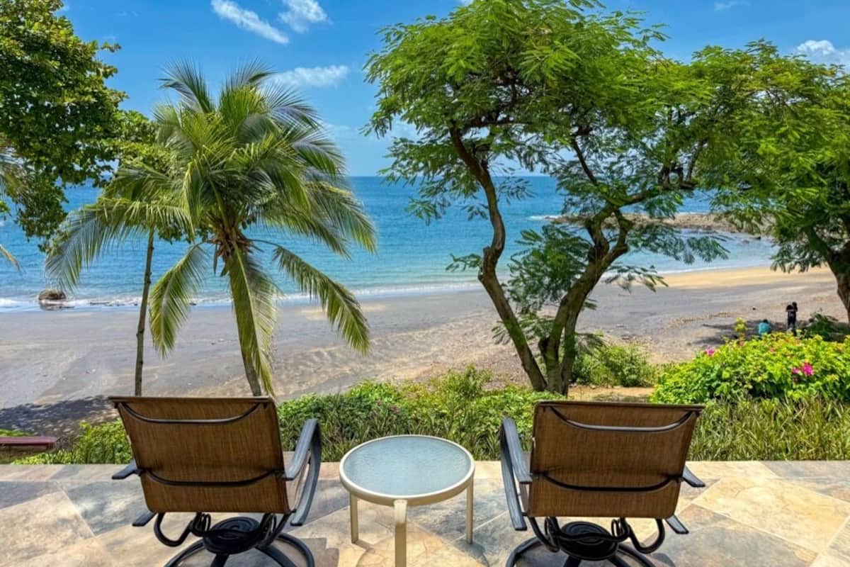 Blick auf einen Strand mit zwei Korbstühlen, einem kleinen Tisch, Palmen und einem klaren, blauen Himmel.
