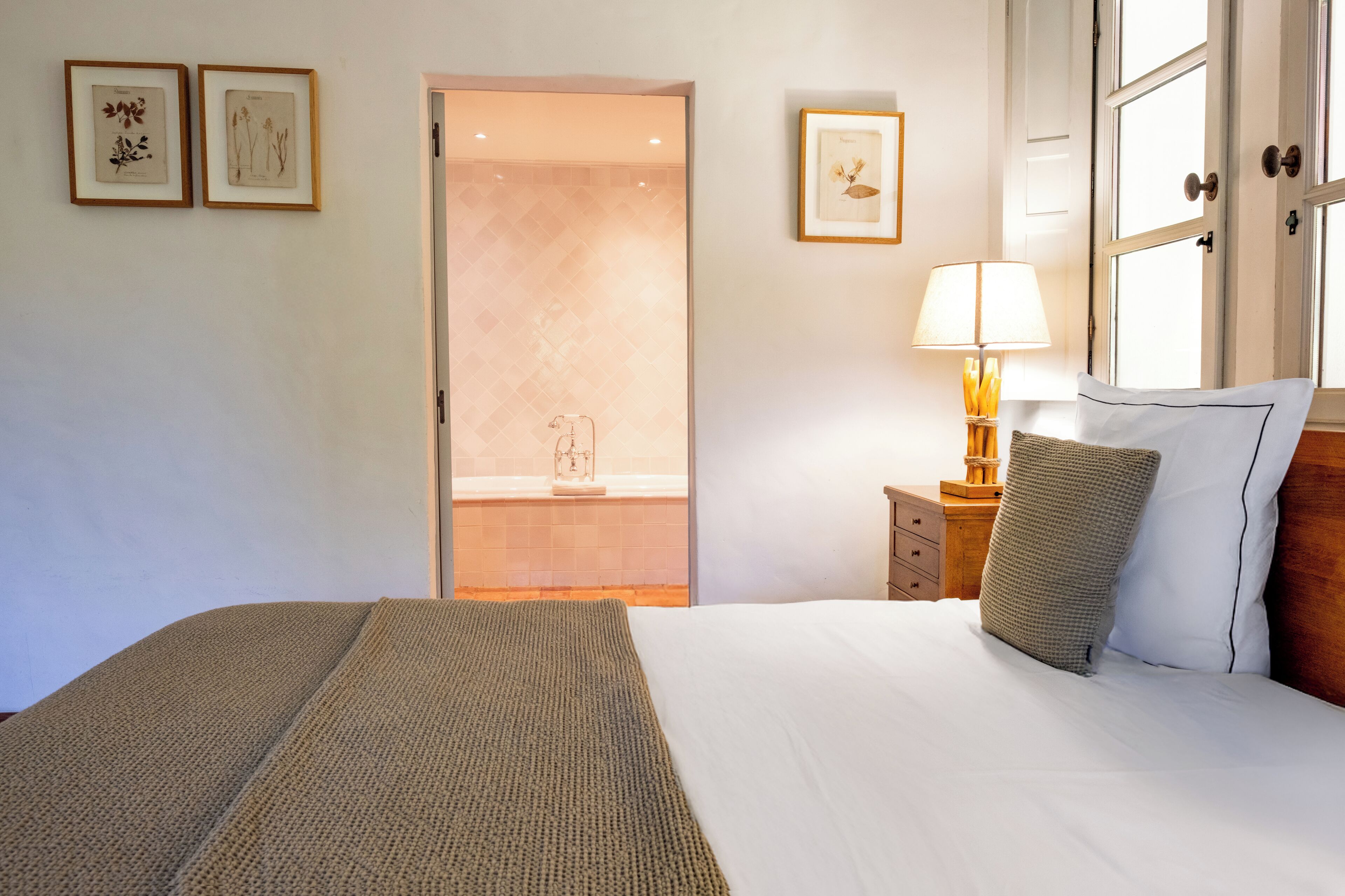 A neatly arranged bedroom with a large bed, a bedside table with a lamp, and framed botanical prints on the wall.