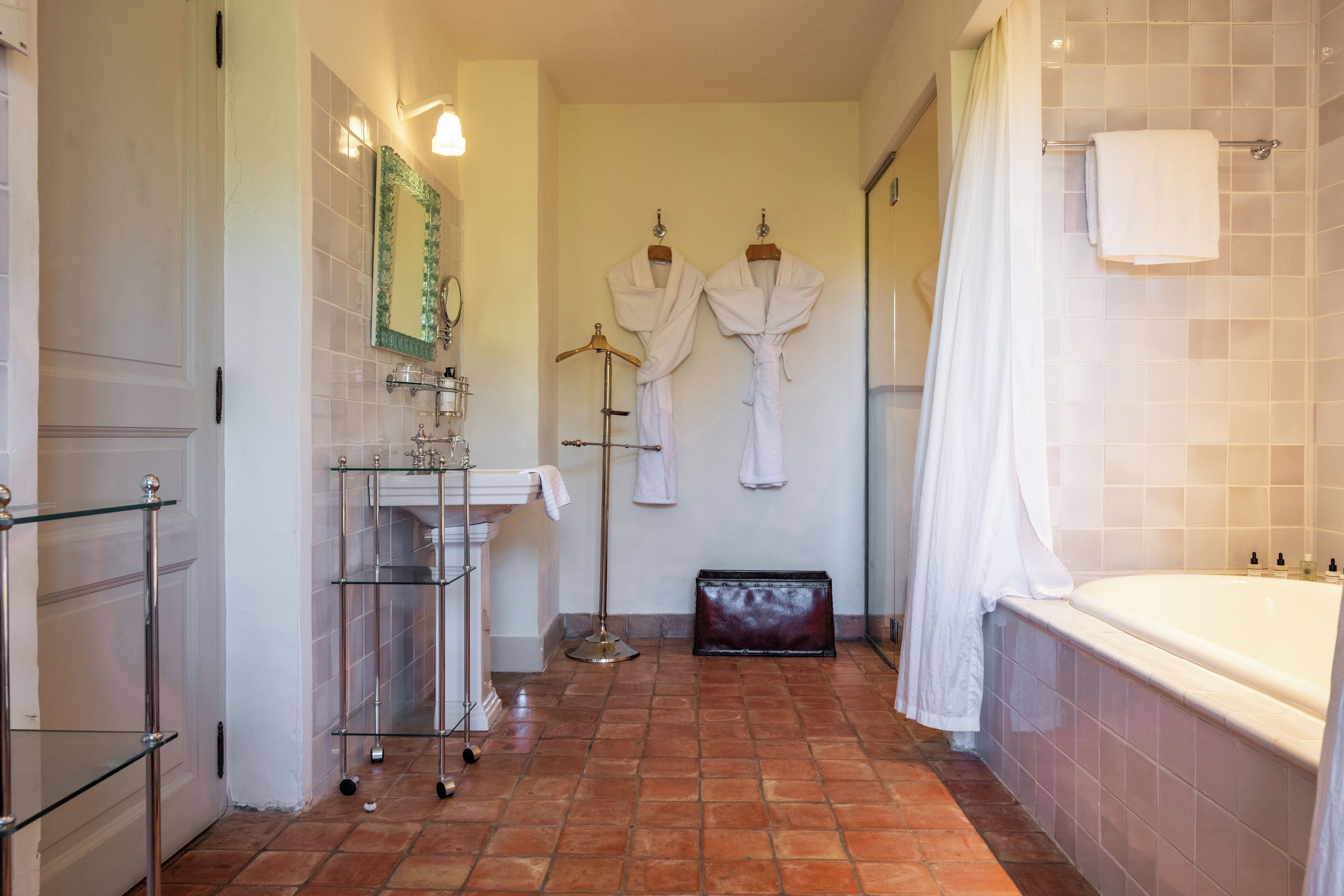 A bathroom with a bathtub, a glass shelf with toiletries, and two white robes hanging on a rack.