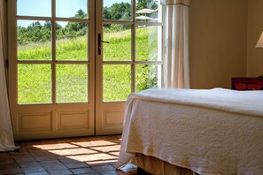 A room with a bed, a window with a view of a green field, and a red piece of furniture.