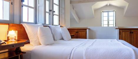 A bedroom with a large bed, wooden headboard, and a nightstand with a lamp. There are multiple windows providing natural light.