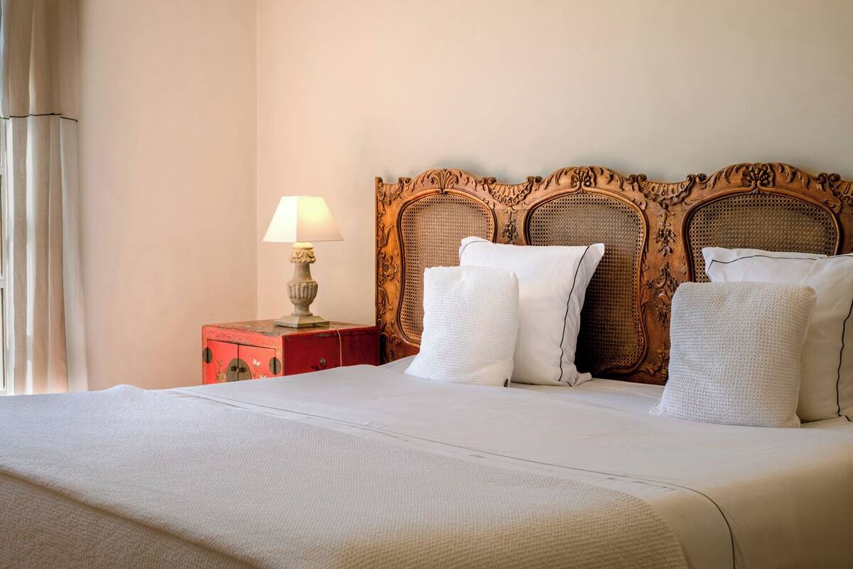 A neatly made bed with white linens, a wooden headboard with intricate carvings, and a small bedside table with a lamp.