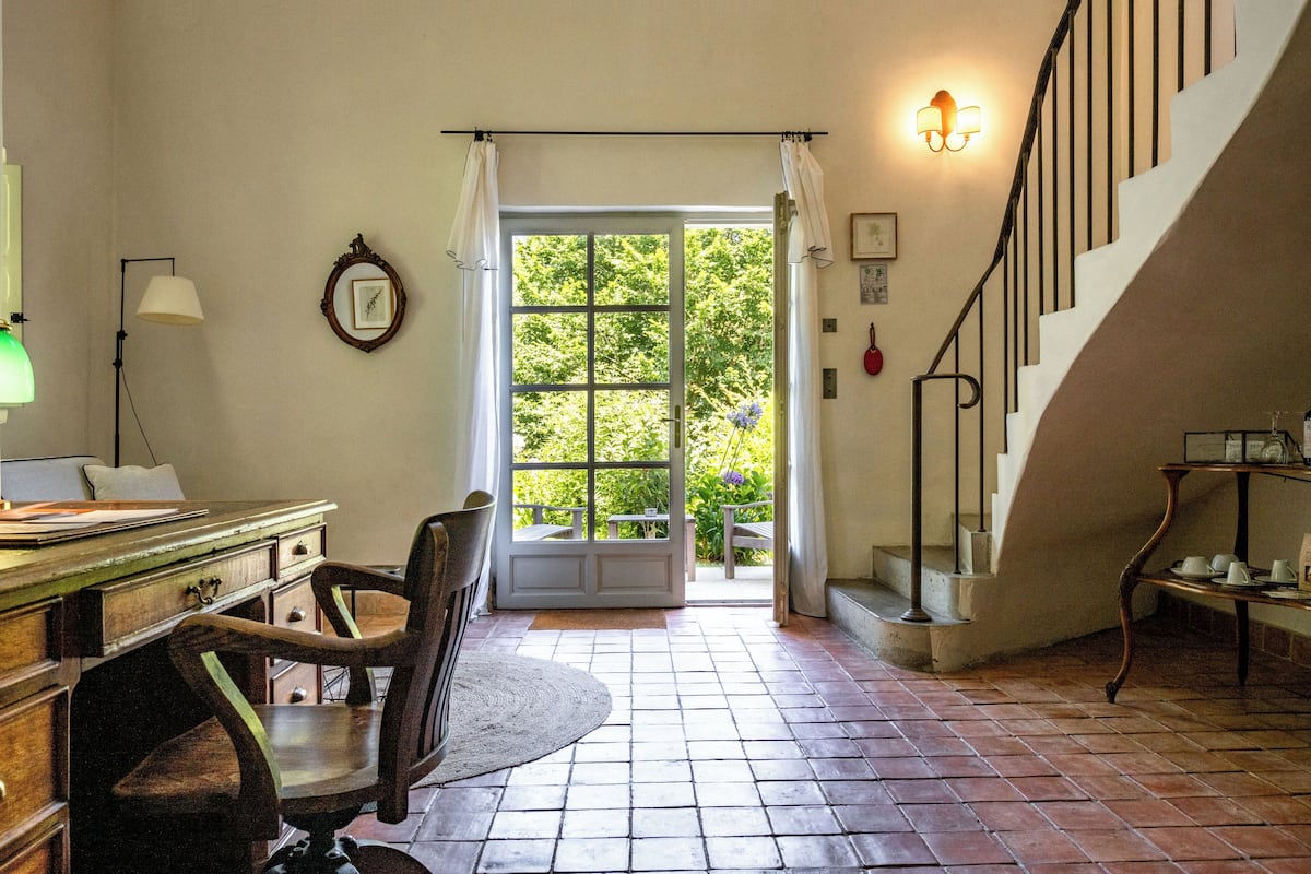 A room with a wooden desk, chair, and a view of a garden through a large window.