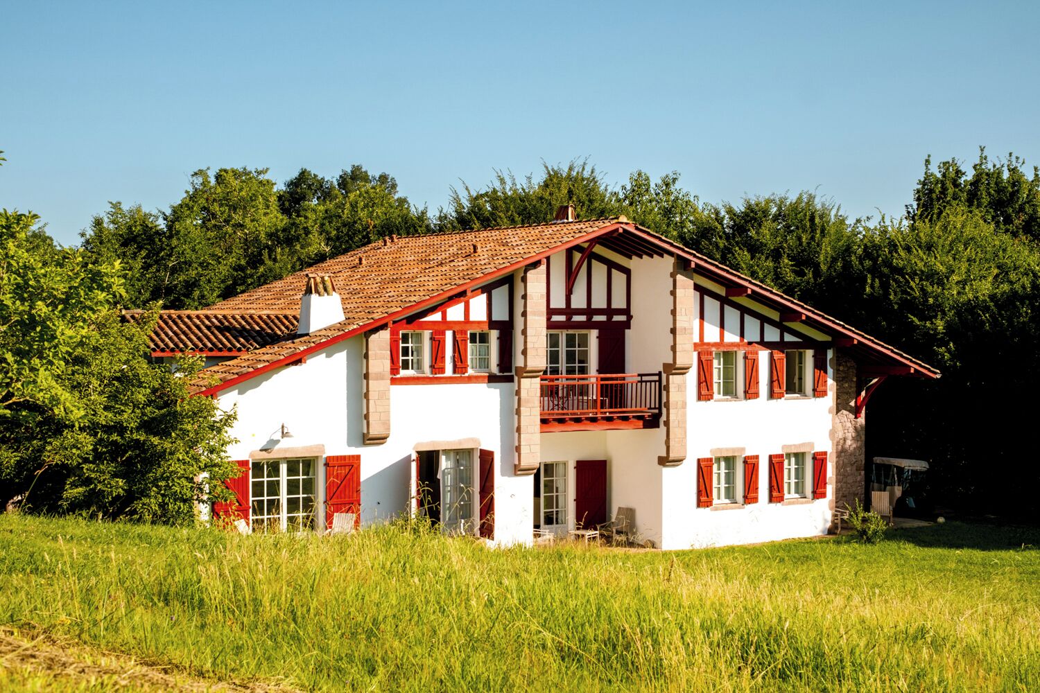 A two-story house with a red-tiled roof, white walls, and red shutters. It has a balcony on the upper floor and is surrounded by greenery.