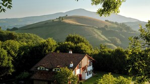 Una casa con techo inclinado, rodeada de abundante vegetación y colinas al fondo.