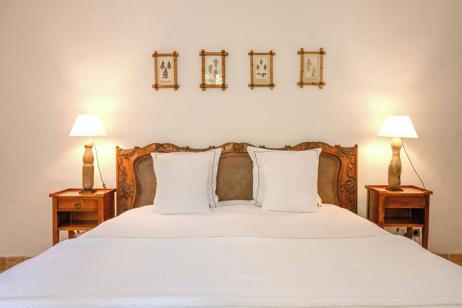 Una cama bien hecha con sábanas blancas, flanqueada por dos mesitas de noche con lámparas, y un cabecero con respaldo tejido.