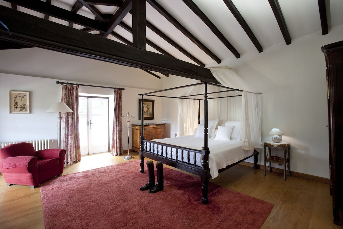 A bedroom with a four-poster bed, a red armchair, a wooden dresser, and a small table with a lamp.