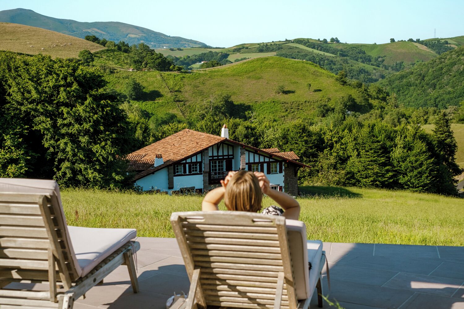 Une vue d’une maison avec un toit en pente et un balcon, entourée de collines verdoyantes et d’arbres.