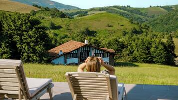 Une vue d’une maison avec un toit en pente et un balcon, entourée de collines verdoyantes et d’arbres.