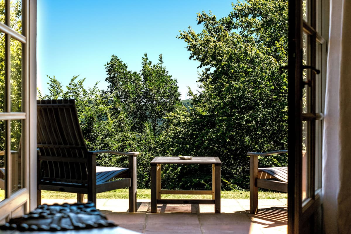 An outdoor patio with a table and two chairs, a view of a lush green garden, and a clear blue sky.