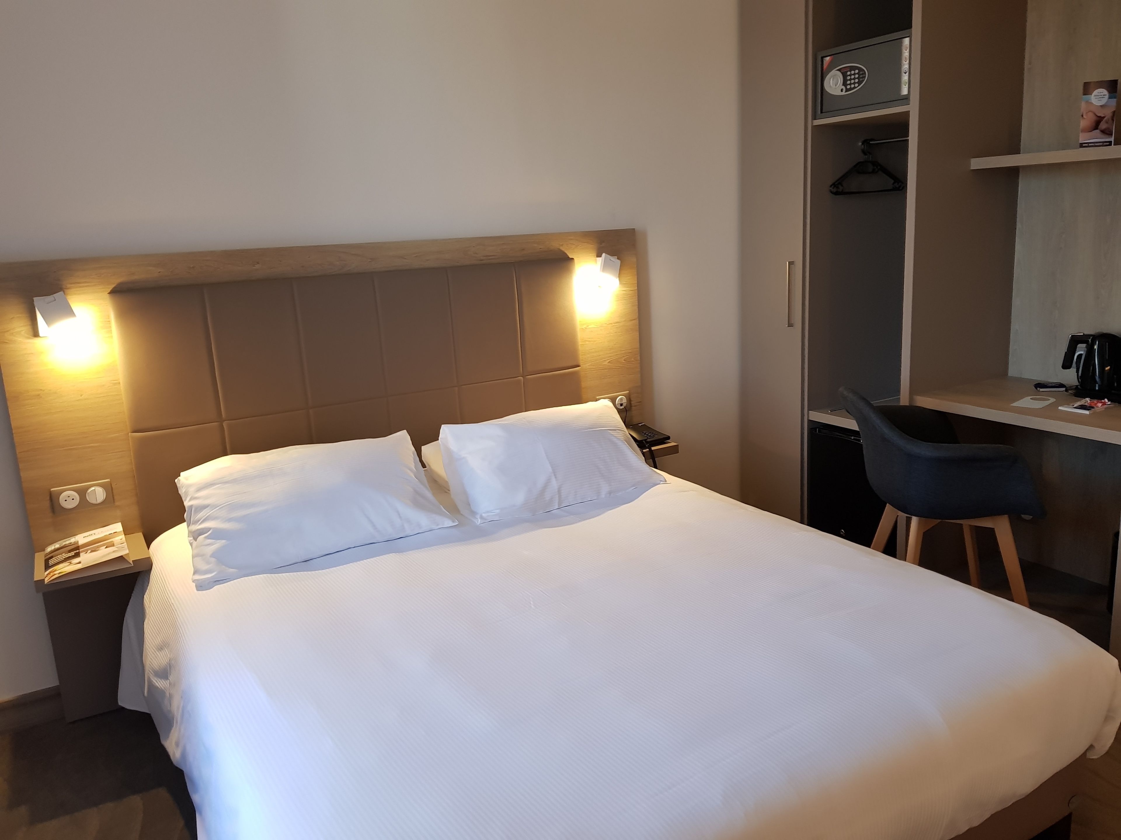 A neatly made bed with white linens, a bedside table with a phone and a lamp, and a desk with a chair and a coffee maker.