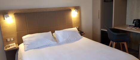 A neatly made bed with white linens, a bedside table with a phone and a lamp, and a desk with a chair and a coffee maker.