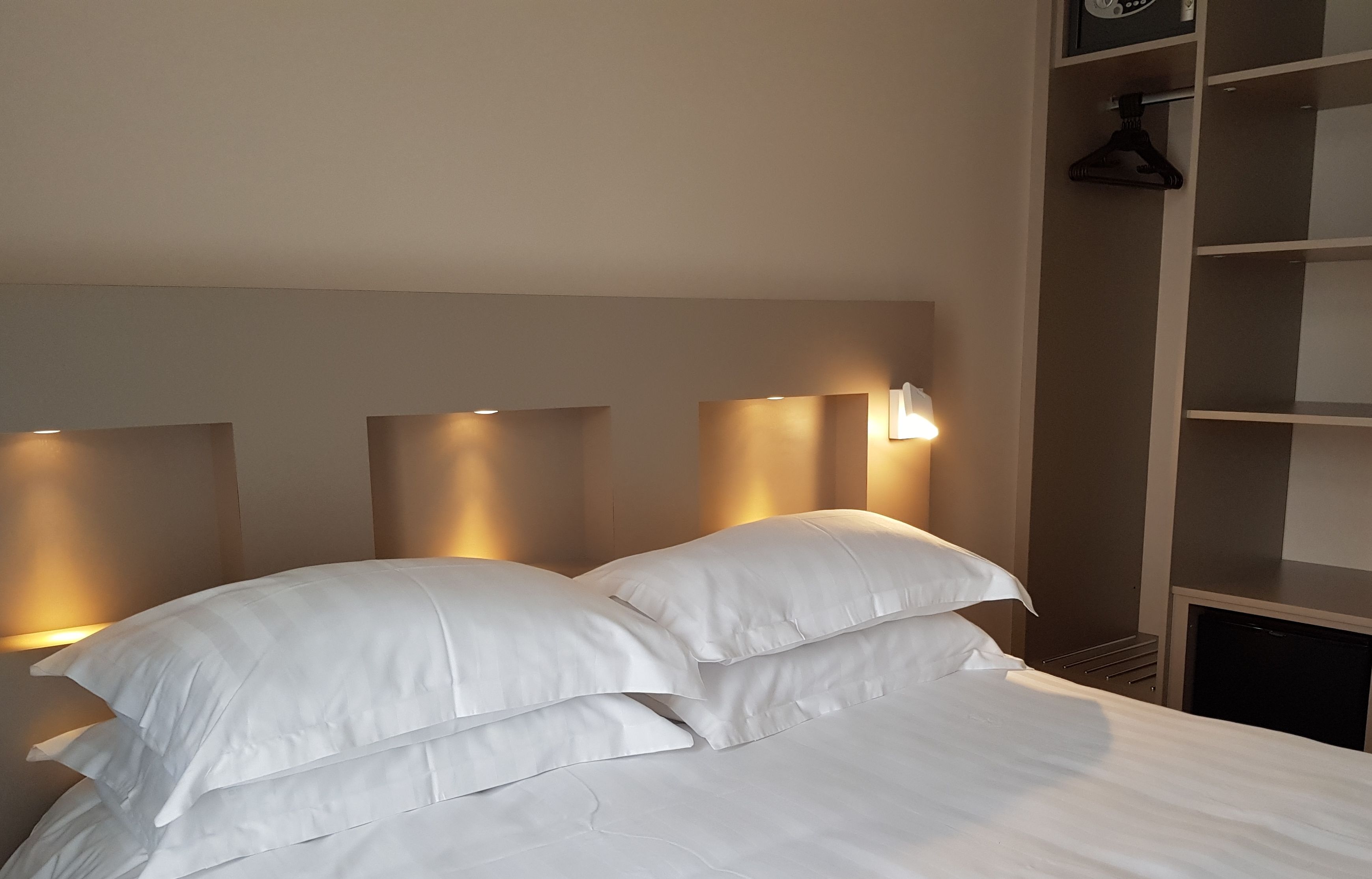 A neatly made bed with white linens and pillows, a wall-mounted light fixture, and a shelving unit with a hanging garment.