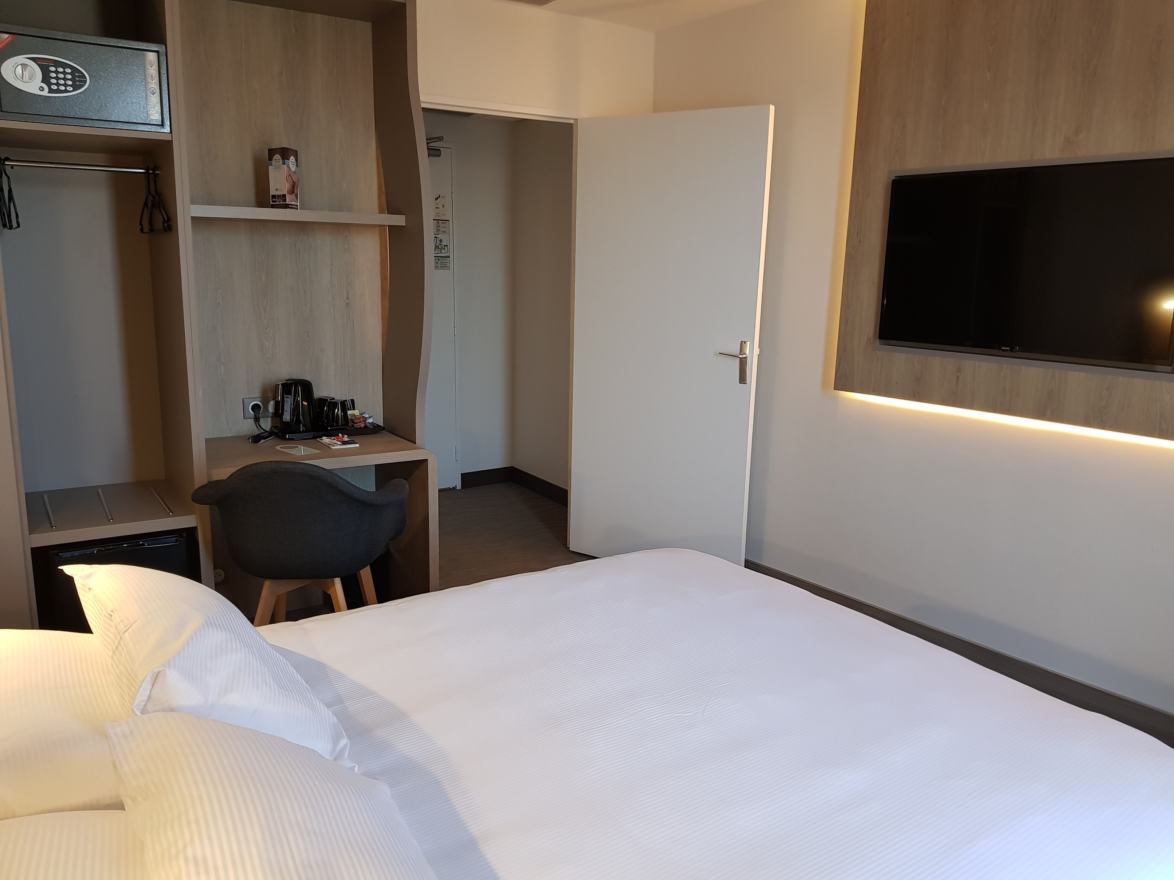 A hotel room with a bed, a desk with a chair, a TV mounted on the wall, and a wardrobe.