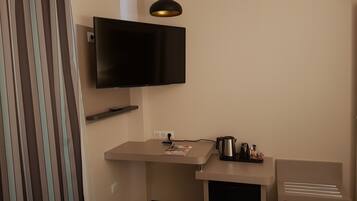 Habitación funcional con televisión de pantalla plana en la pared, una pequeña zona de kitchenette con cafetera y una silla blanca.