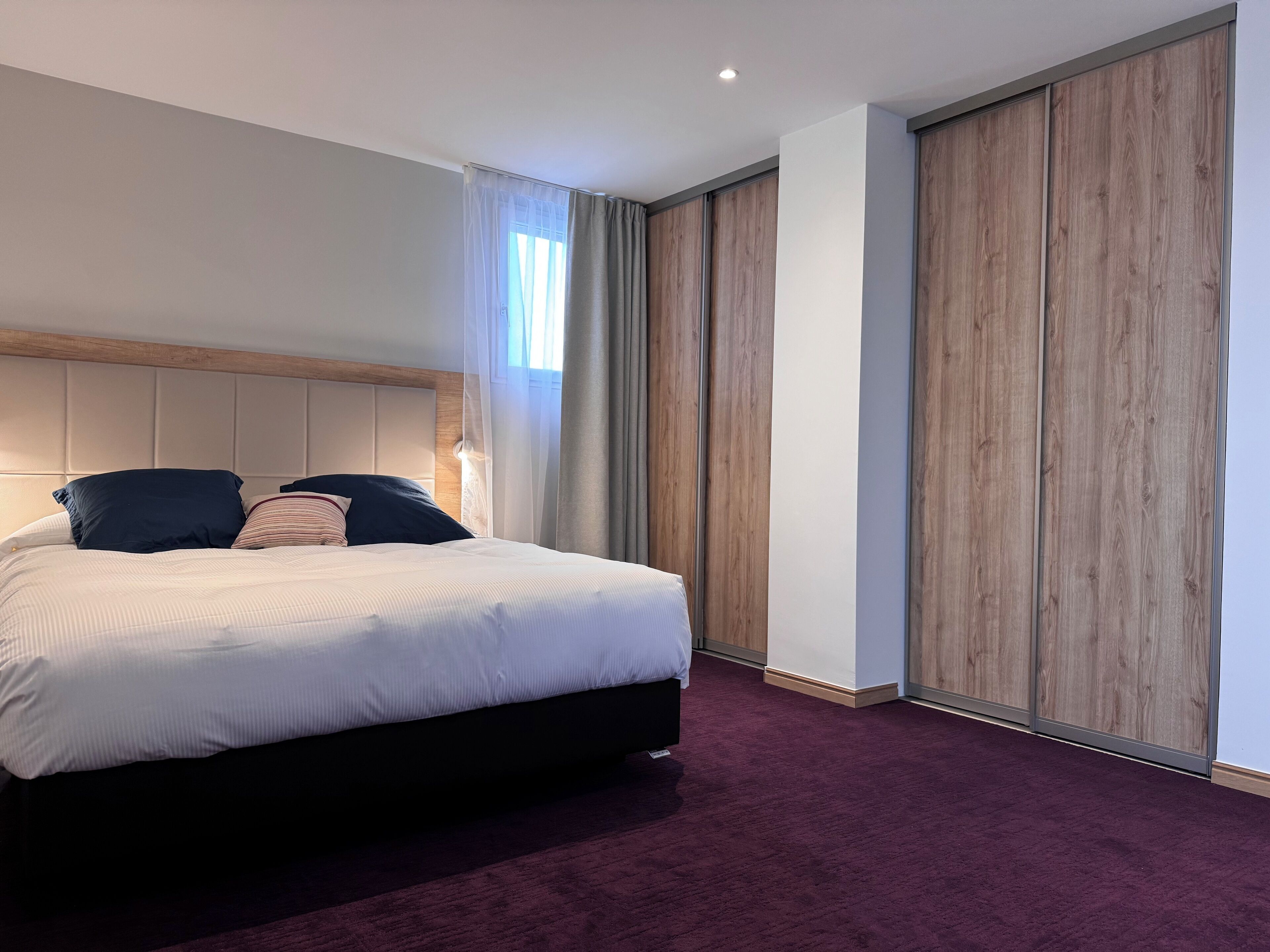 Une chambre d’hôtel moderne avec un grand lit, une tête de lit en bois, une moquette sombre et une grande fenêtre avec des rideaux.