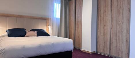 Une chambre d’hôtel moderne avec un grand lit, une tête de lit en bois, une moquette sombre et une grande fenêtre avec des rideaux.