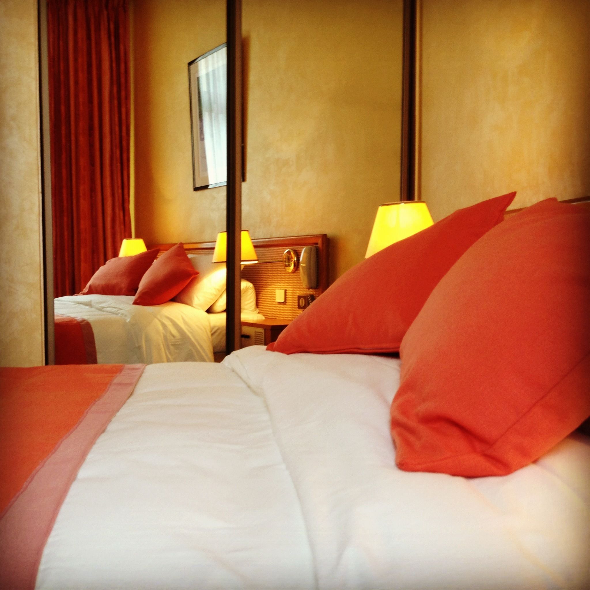 A hotel room with two beds, each with white linens and red pillows. There are two bedside lamps and a wooden headboard.