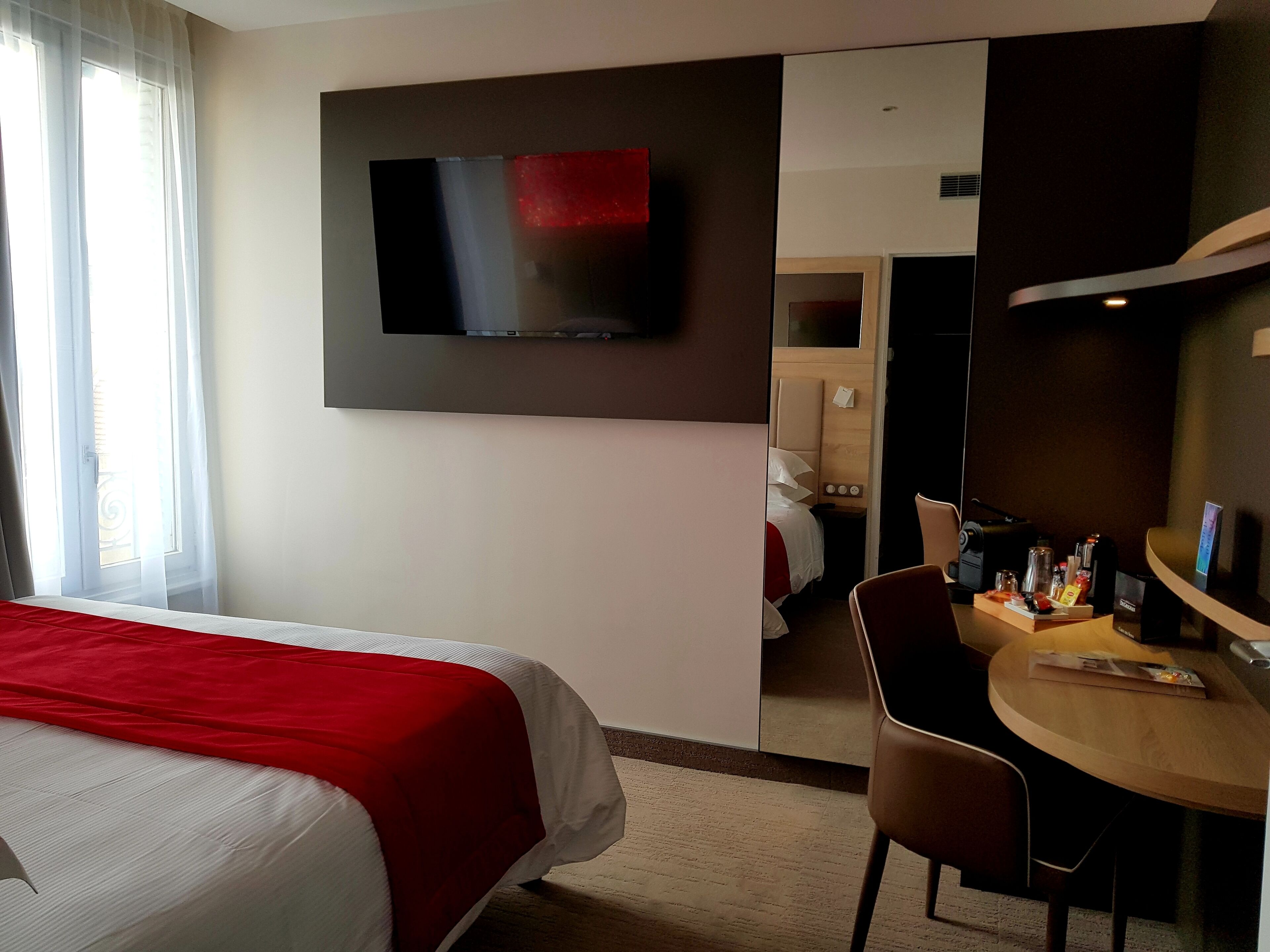 A hotel room with a bed, a TV mounted on the wall, a desk with a chair, and a small table with various items.