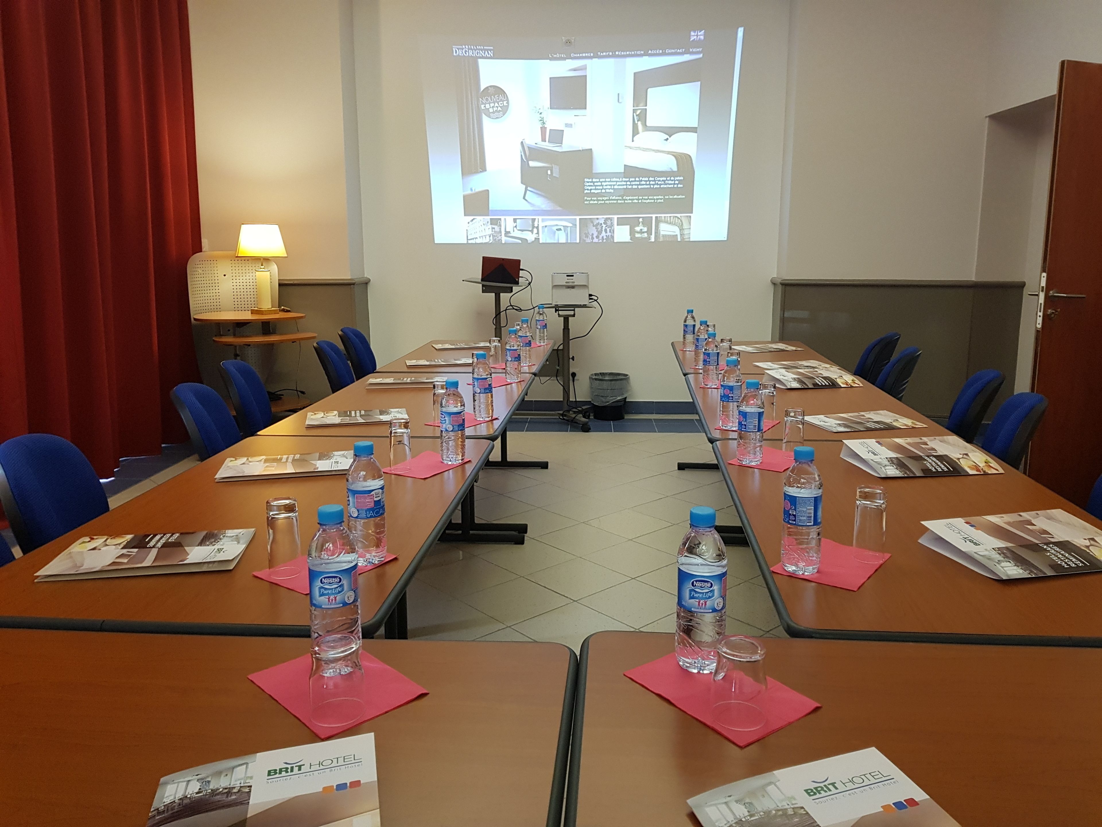 A conference room with a long table, blue chairs, water bottles, and a projector screen displaying a hotel room.