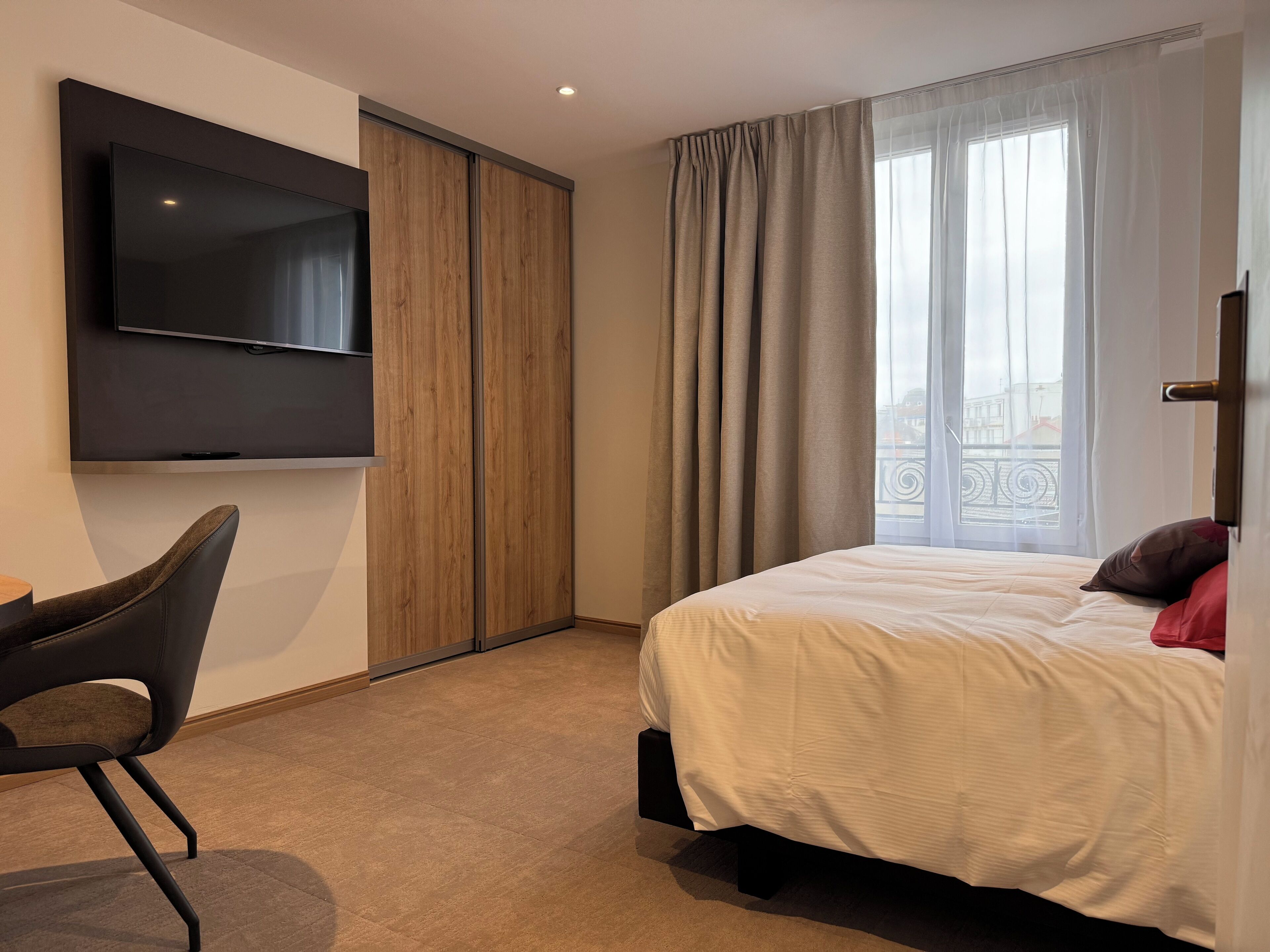 Een moderne hotelkamer met een bed, een aan de muur bevestigde televisie, een stoel en uitzicht op de stad door het raam.