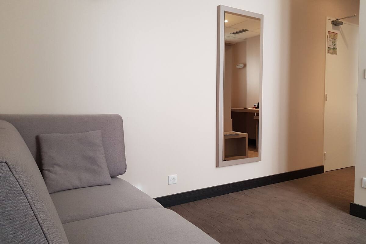 A modern living room with a grey sofa, a large mirror, and a hallway leading to a bathroom.