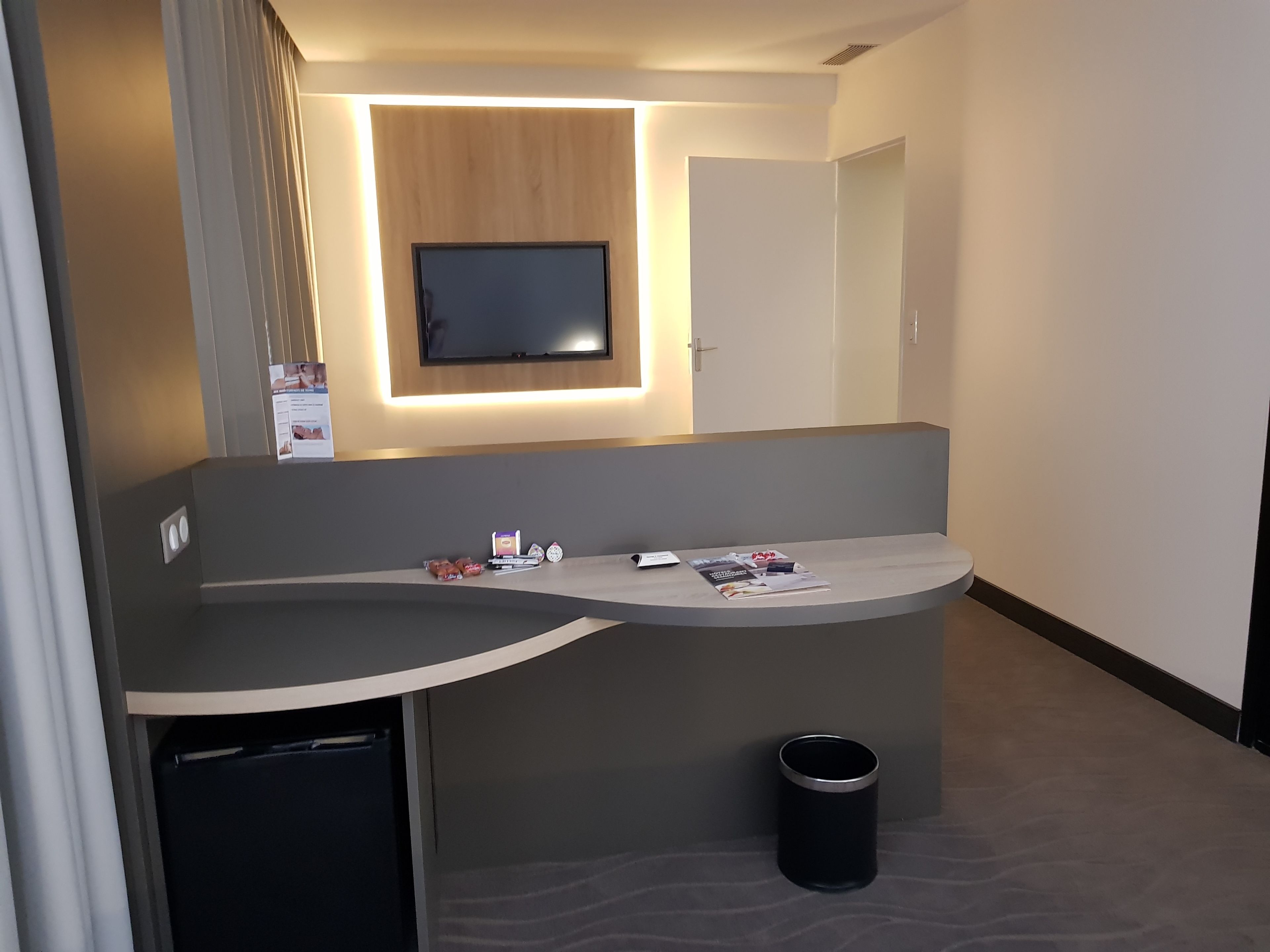 A modern hotel room with a curved desk, a flat-screen TV mounted on the wall, a small trash bin, and a door.