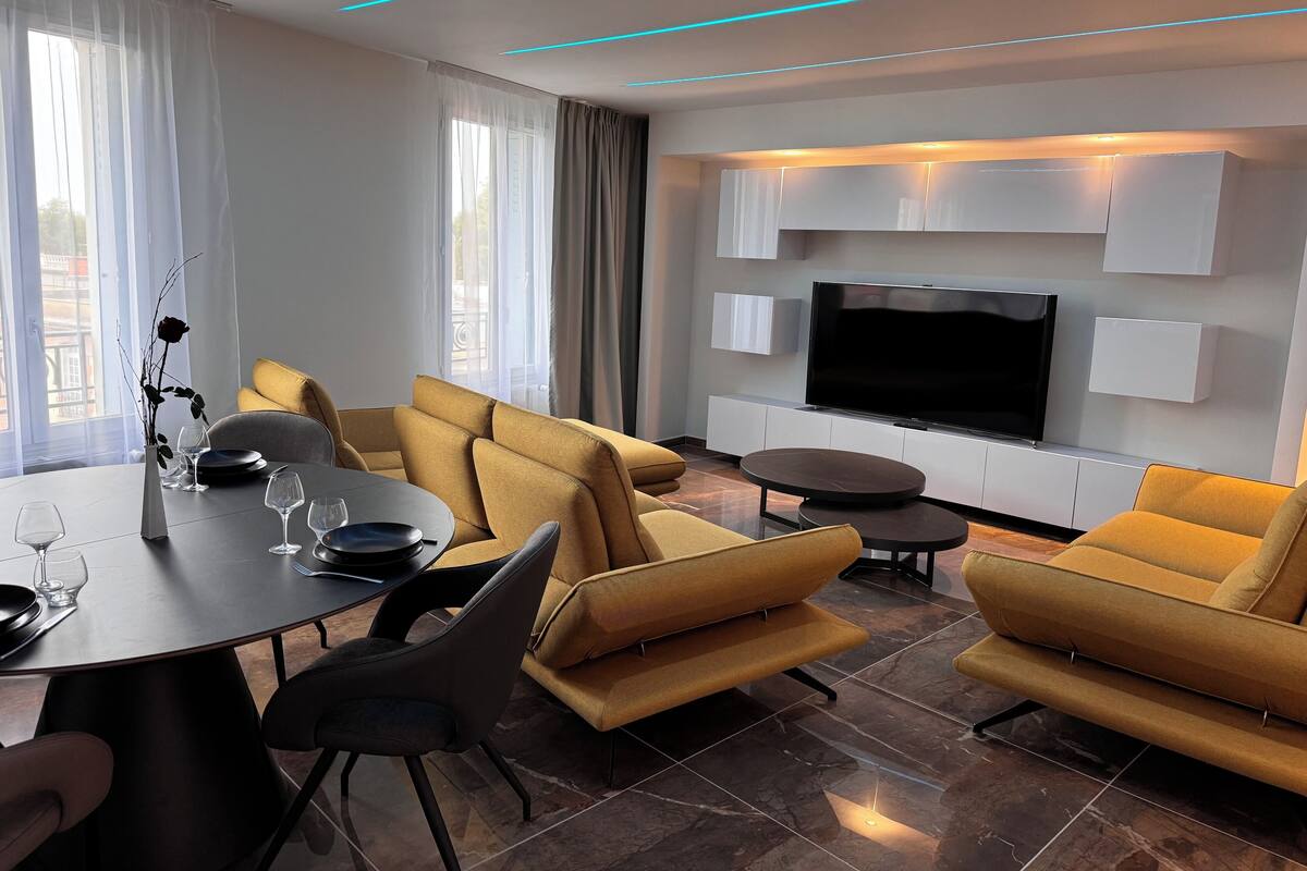 A modern living room with a dining area, featuring a sofa set, a flat-screen TV, and a dining table with wine glasses.