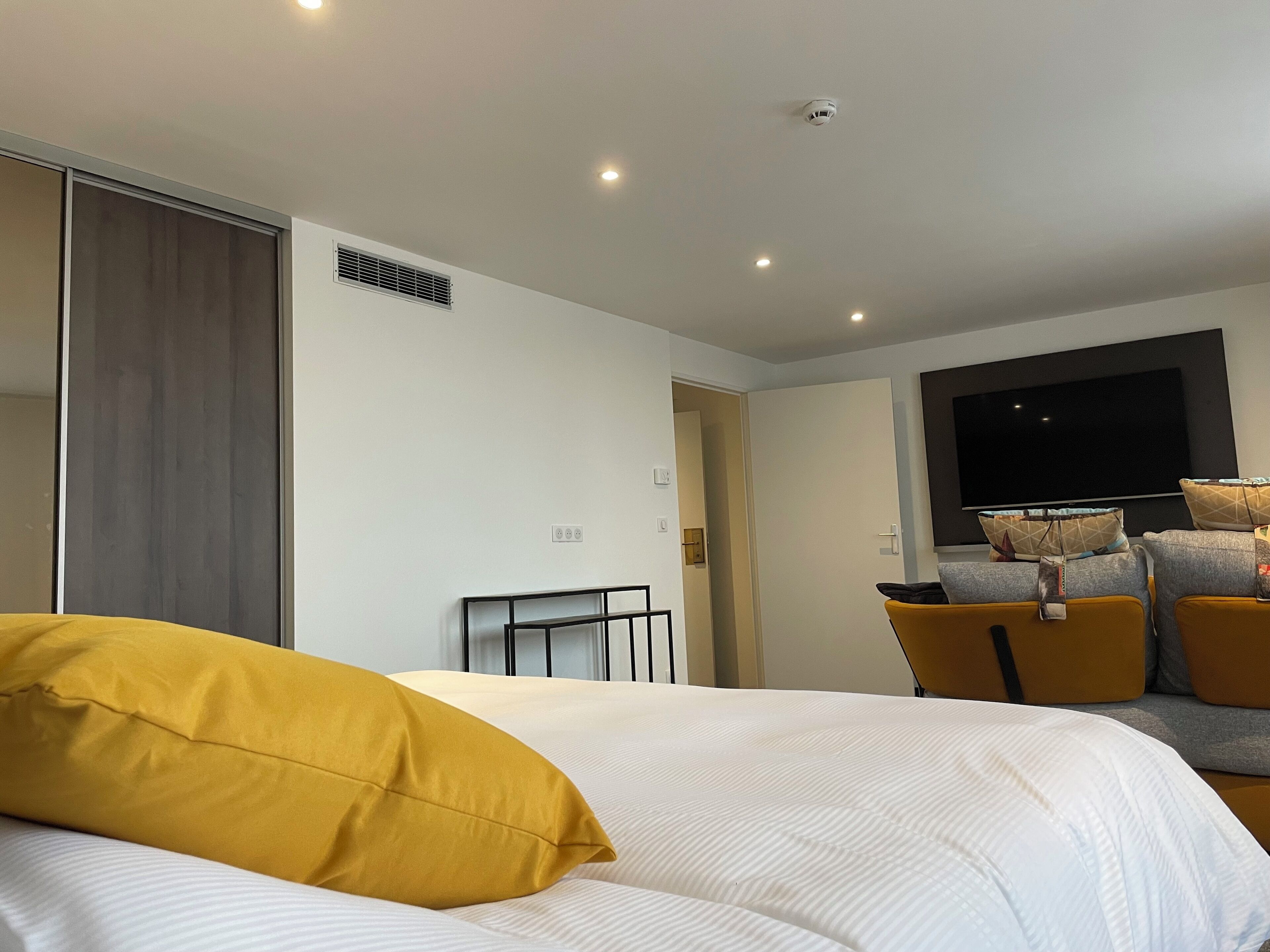 A modern hotel room with a large bed, a sofa, a TV, and a ceiling-mounted smoke detector.