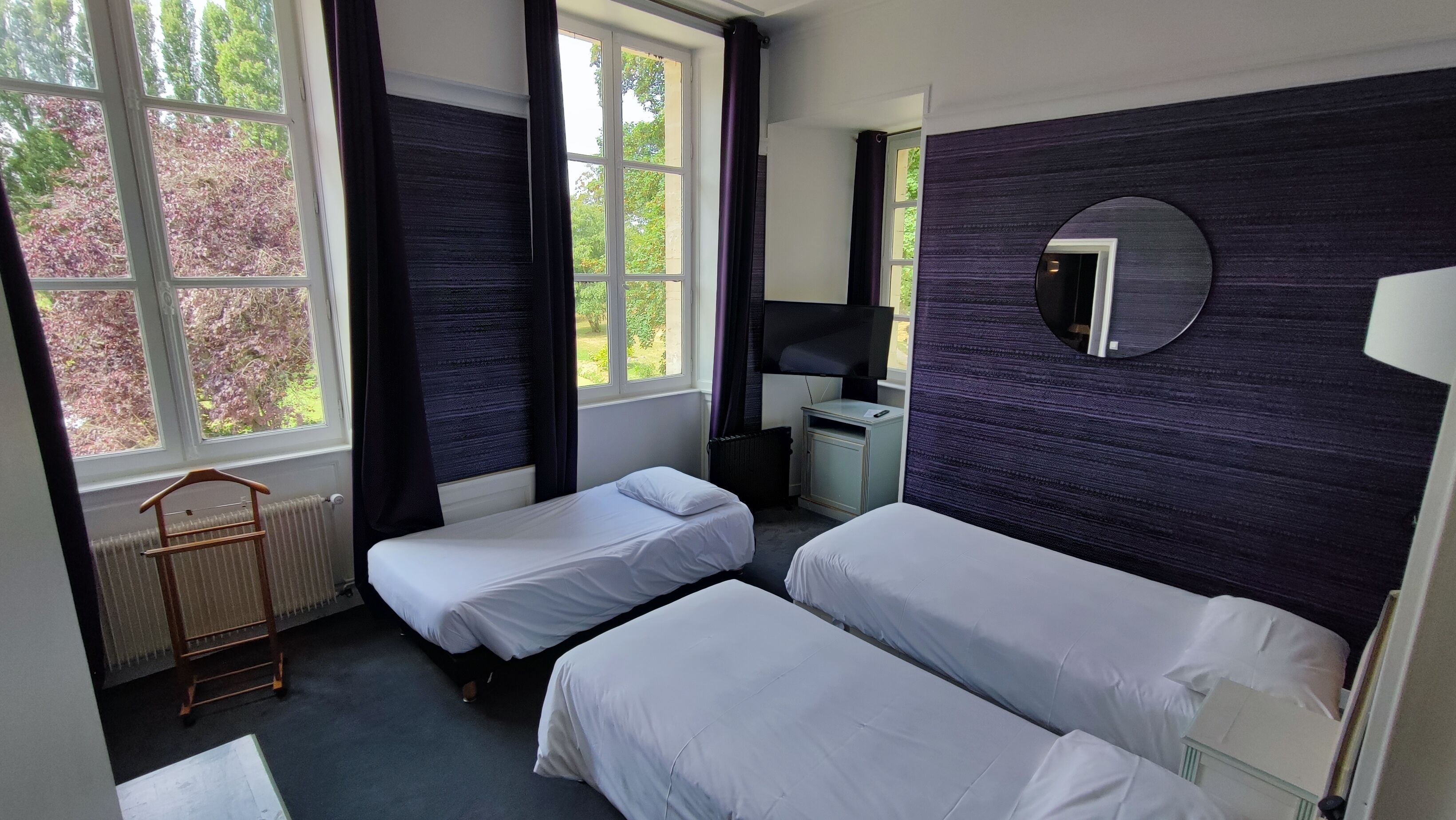 A hotel room with two single beds, a small desk, a chair, a radiator, and a window with curtains.