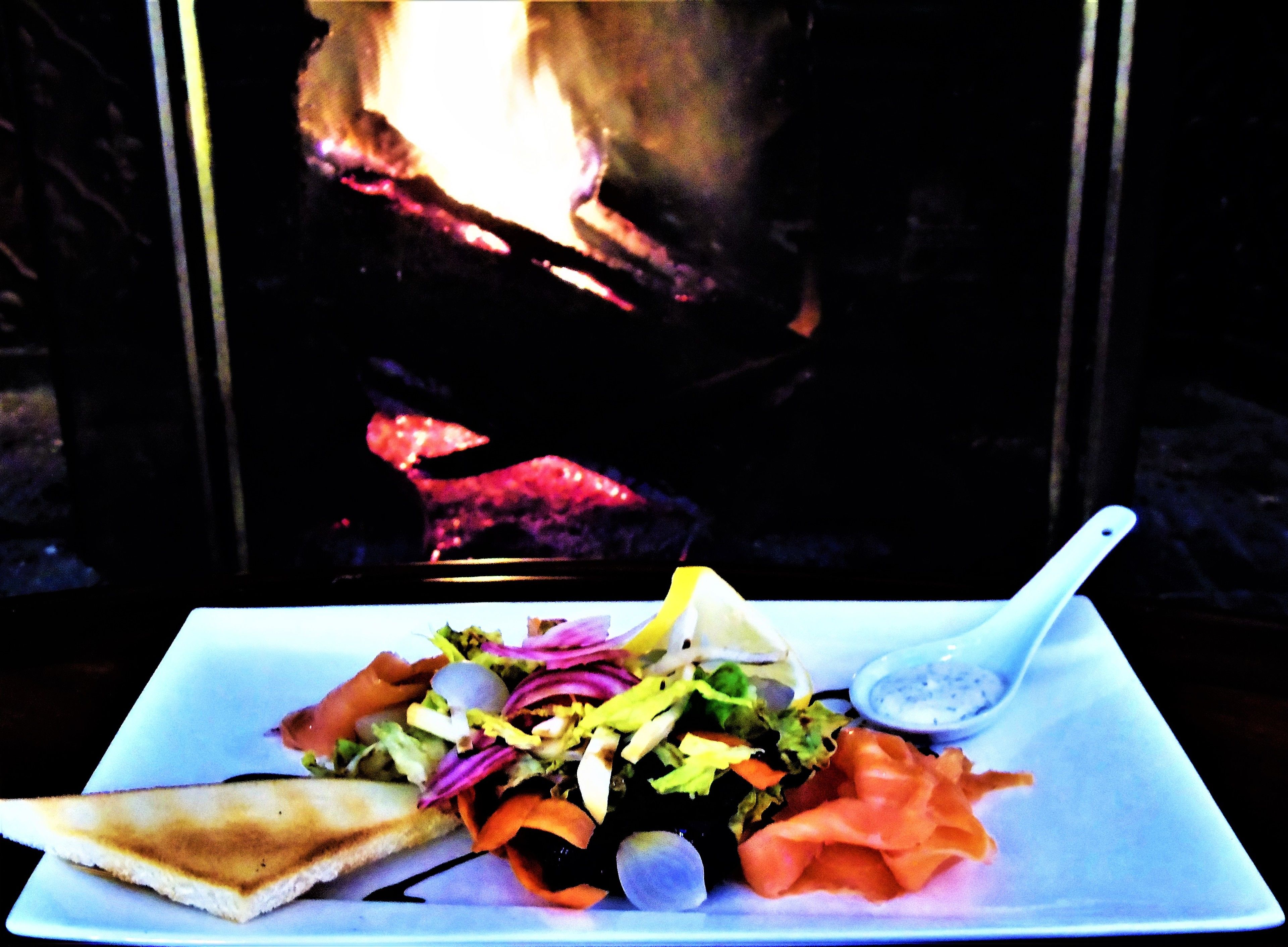 En tallrik med mat och en vit sked, innehållande en smörgås, en sallad och rökt lax, placerad framför en öppen spis.