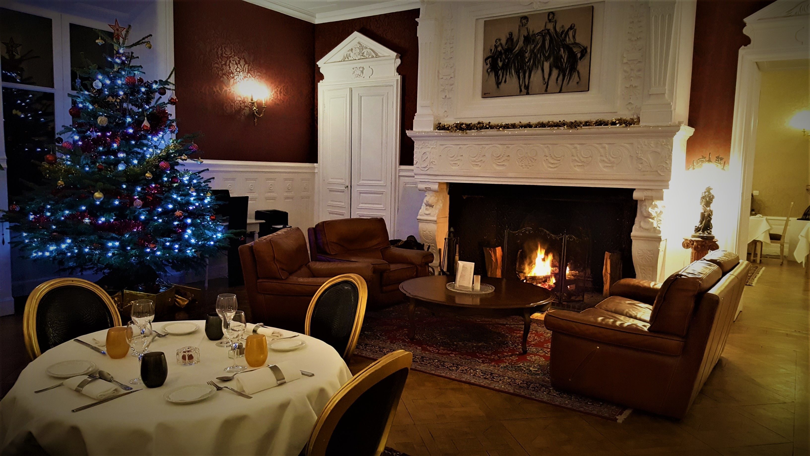 A warmly lit living room with a fireplace, a Christmas tree, and a dining area set for a meal.