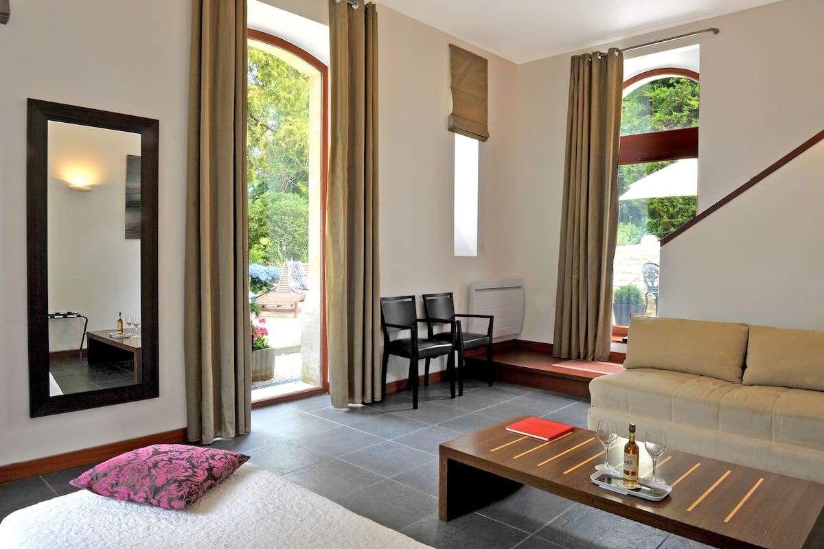 Una habitación de hotel con cama, sofá, una mesita de centro con vasos y una botella, espejo y vistas al exterior.