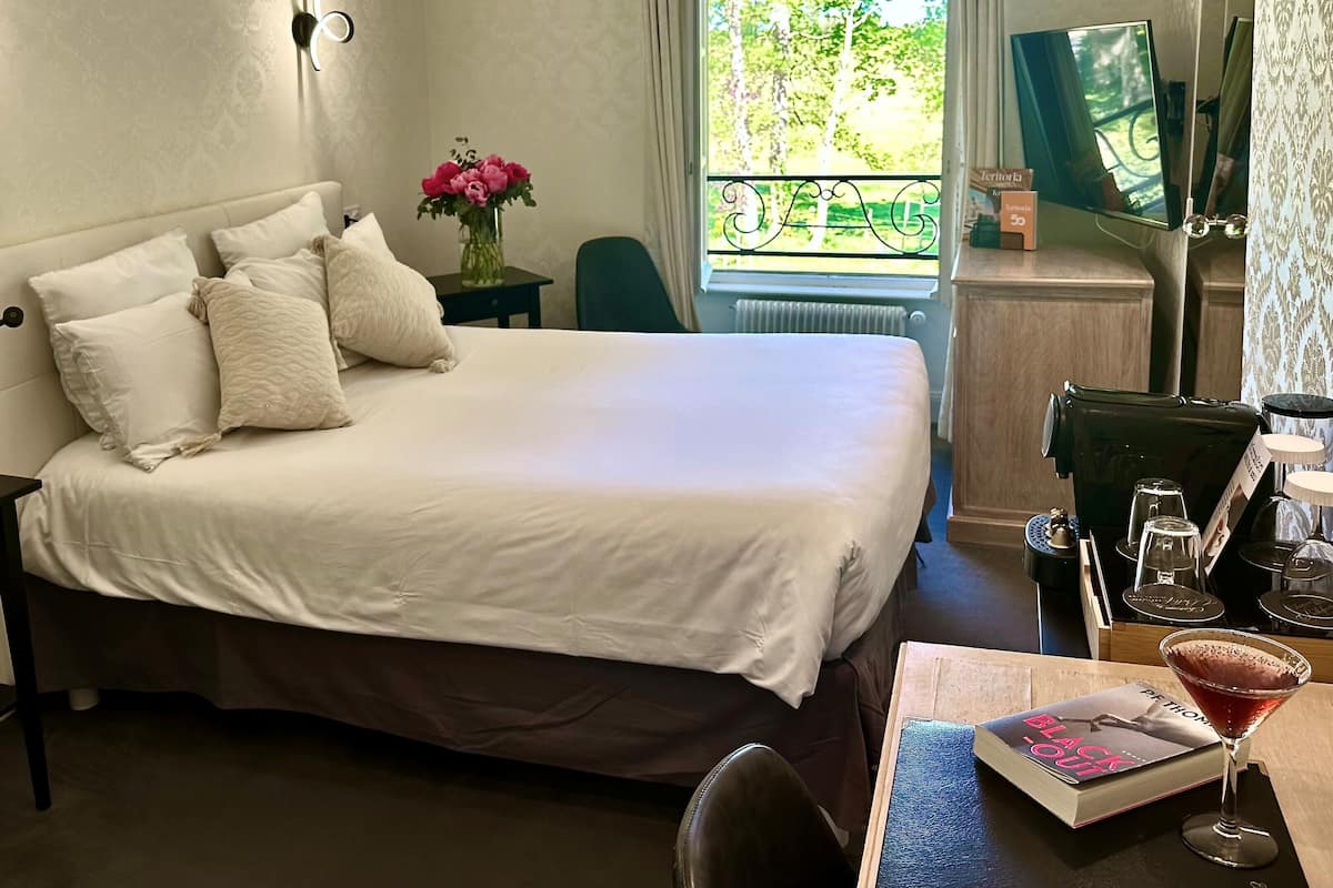 Una cama bien hecha con sábanas blancas y varios almohadones, una mesita con un jarrón de flores, un televisor sobre un mueble bajo y una ventana con vistas a árboles.