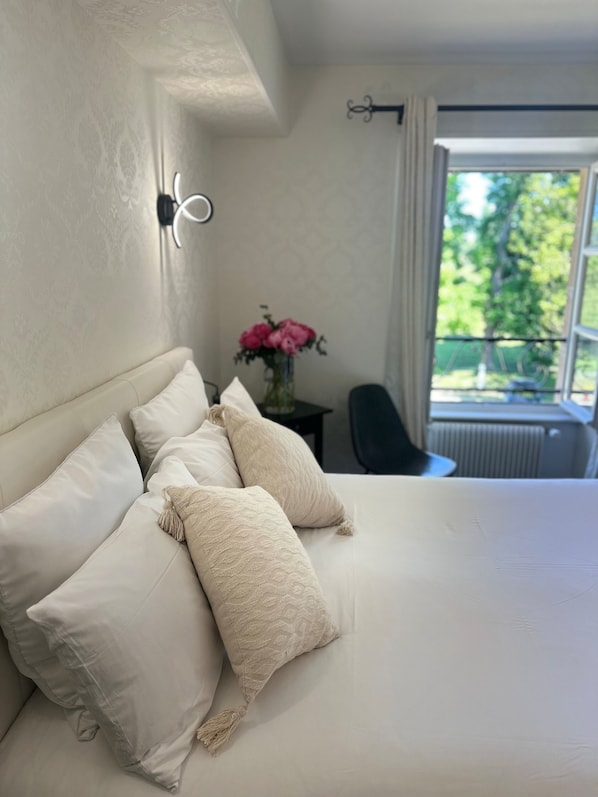 A bedroom with a large bed, white bedding, a vase with pink flowers, and a view of greenery outside.