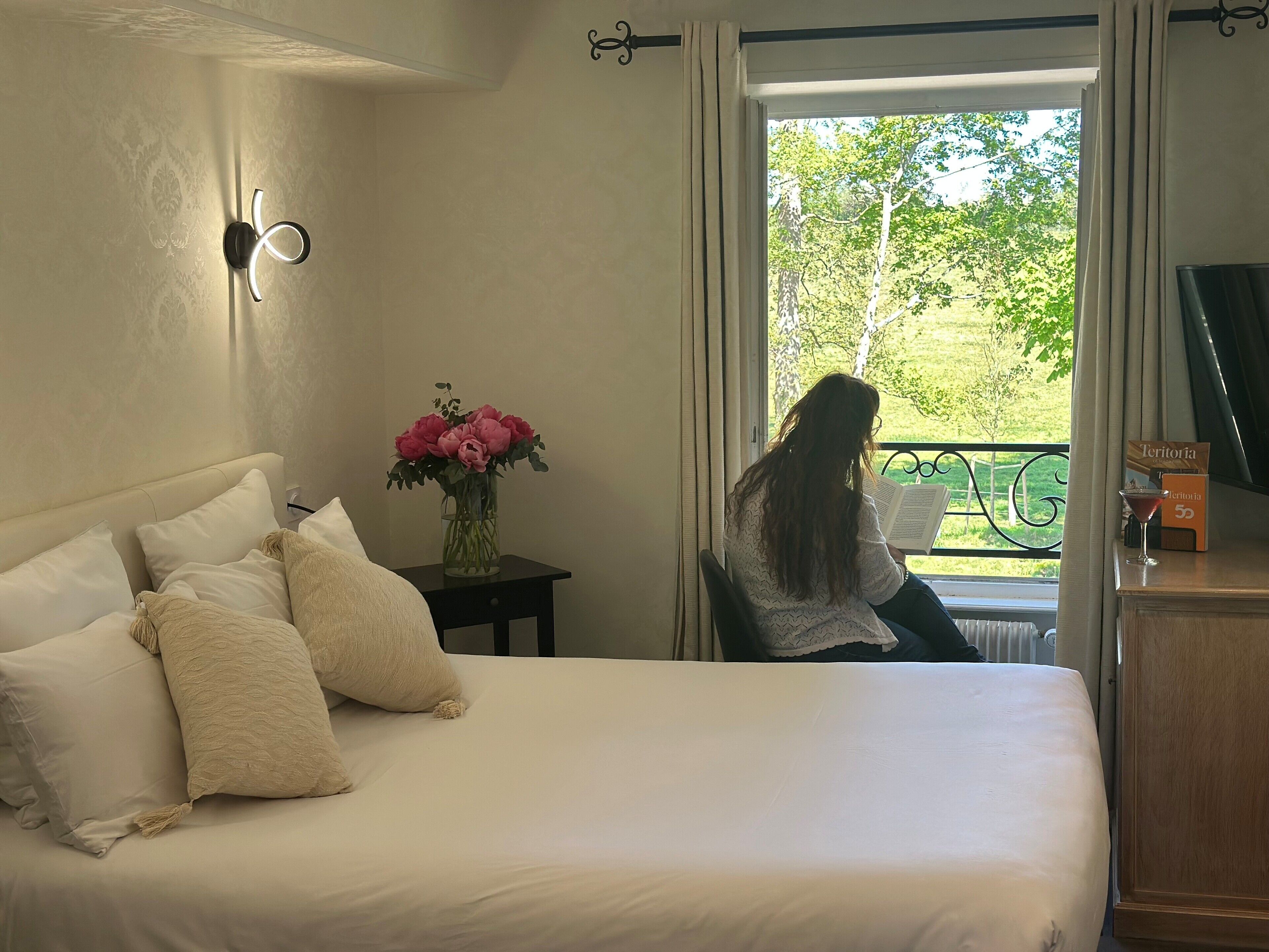 A bedroom with a bed, a nightstand with a vase of flowers, and a view of a balcony with greenery outside.