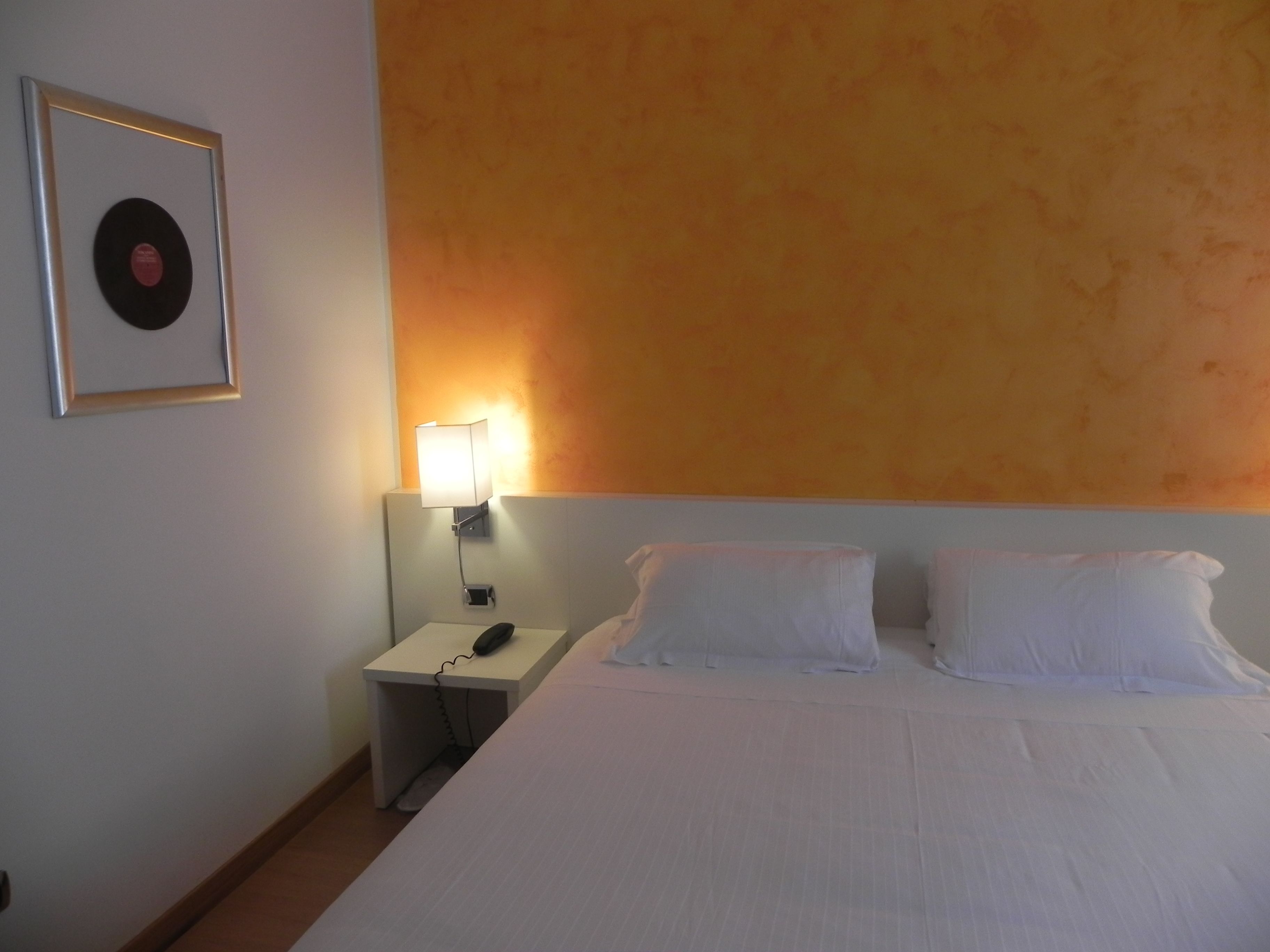Ein Hotelzimmer mit einem Bett, zwei Kissen, einem Nachttisch mit Lampe und einer gerahmten Schallplatte an der Wand.