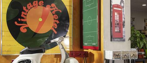 A vintage-themed hotel interior with a scooter, a large poster, a green chalkboard, and a red door illustration.