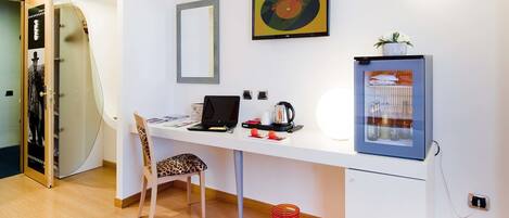 Una habitación moderna con un escritorio blanco, una computadora portátil, una cafetera, un refrigerador, un sillón estampado de leopardo y una puerta de vidrio que da a la zona de la ducha.