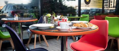 Uma área de refeições com mesa de vidro, cadeiras coloridas e um prato de croissants.