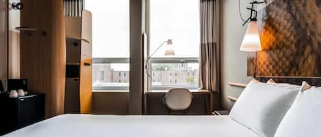 A hotel room with a large bed, a desk with a chair, a view of buildings through the window, and a wall-mounted lamp.